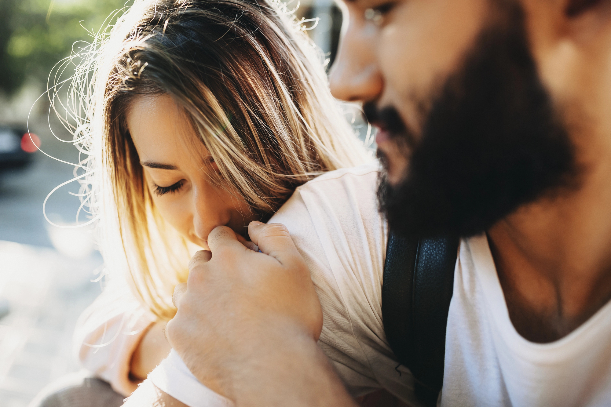 A woman with long, blonde hair gently kisses the shoulder of a bearded man who is holding her close. They both appear to be outdoors, sharing an intimate and affectionate moment. The background is slightly blurred, emphasizing their connection.