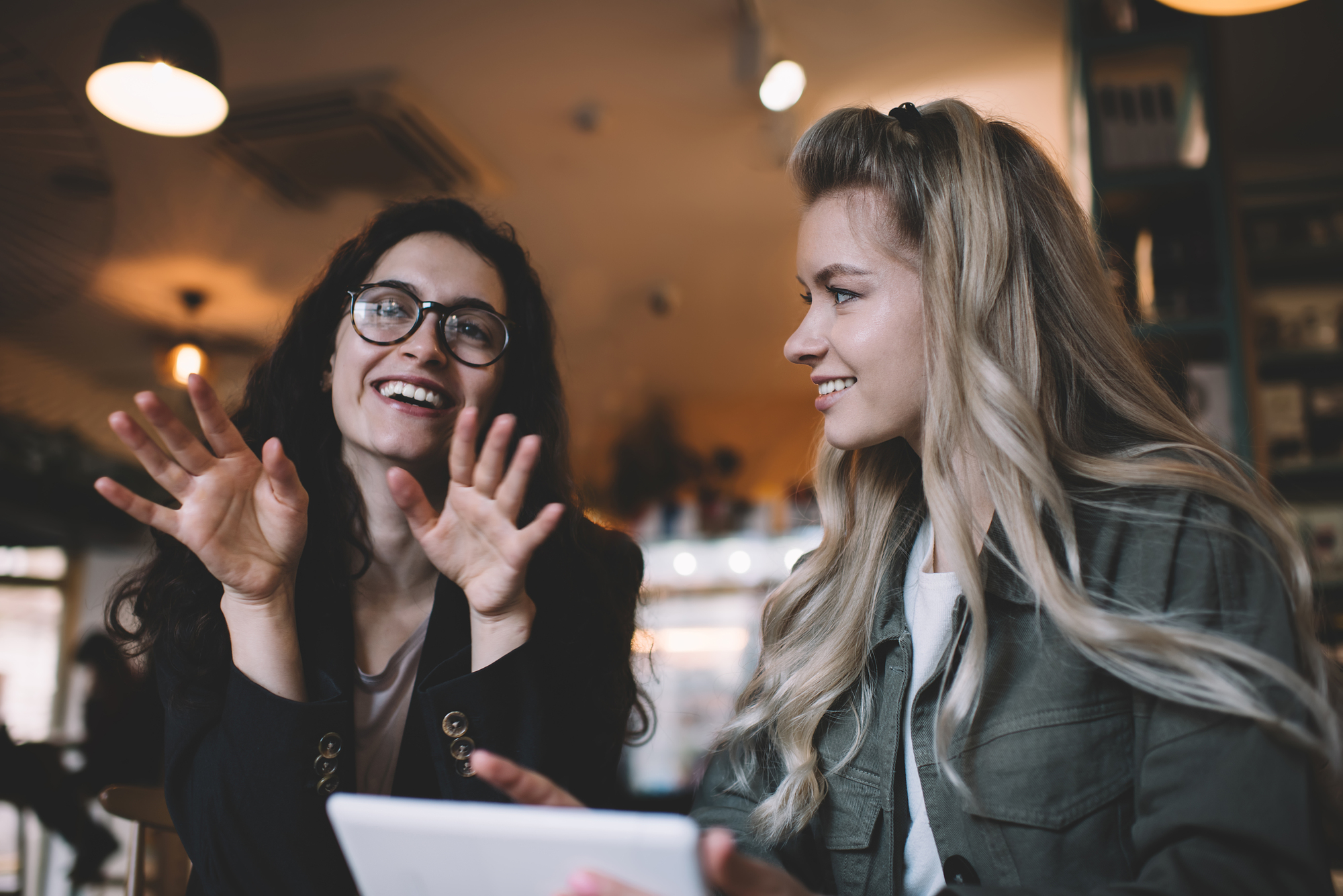 Two women are seated in a cozy café, engaged in a lively conversation. The woman on the left, with curly hair and glasses, is gesturing animatedly with her hands. The woman on the right, with long, blonde hair, is smiling and holding a tablet.