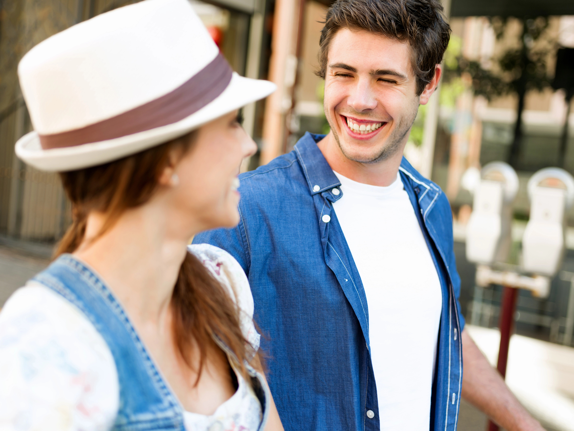 Two people are outdoors, engaging in a lively conversation. The person on the right, wearing a blue shirt and a white t-shirt underneath, is smiling broadly. The person on the left is wearing a white hat and a casual outfit, partially facing the other person.