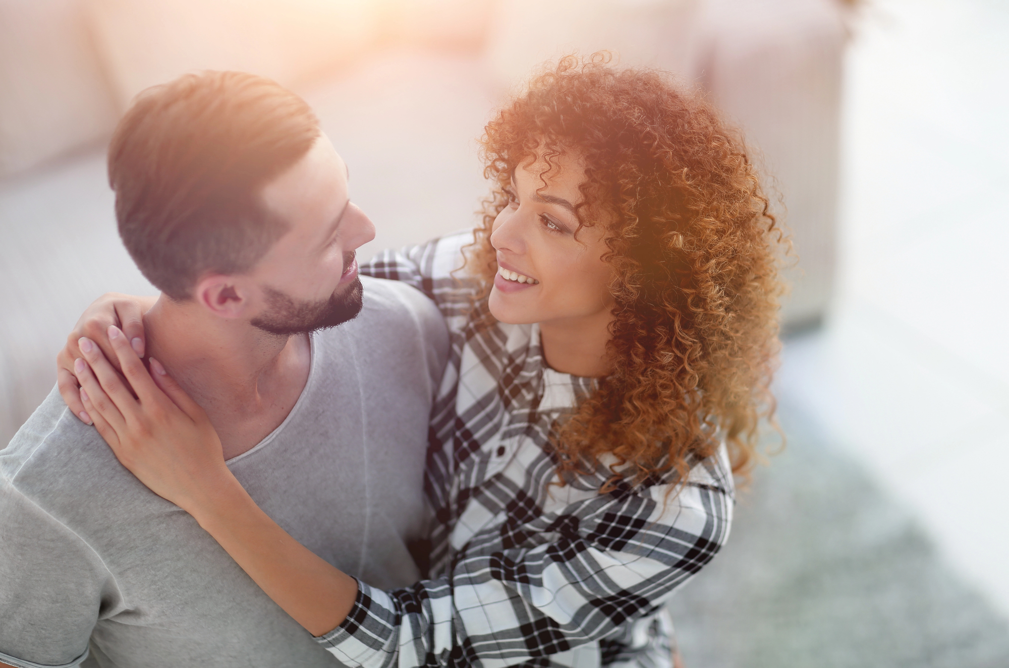 A man and a woman are embracing and looking at each other warmly. The woman, with curly hair and wearing a plaid shirt, has her arms around the man's shoulders. The man, with short hair and a beard, is wearing a light-colored shirt. The background is softly blurred.