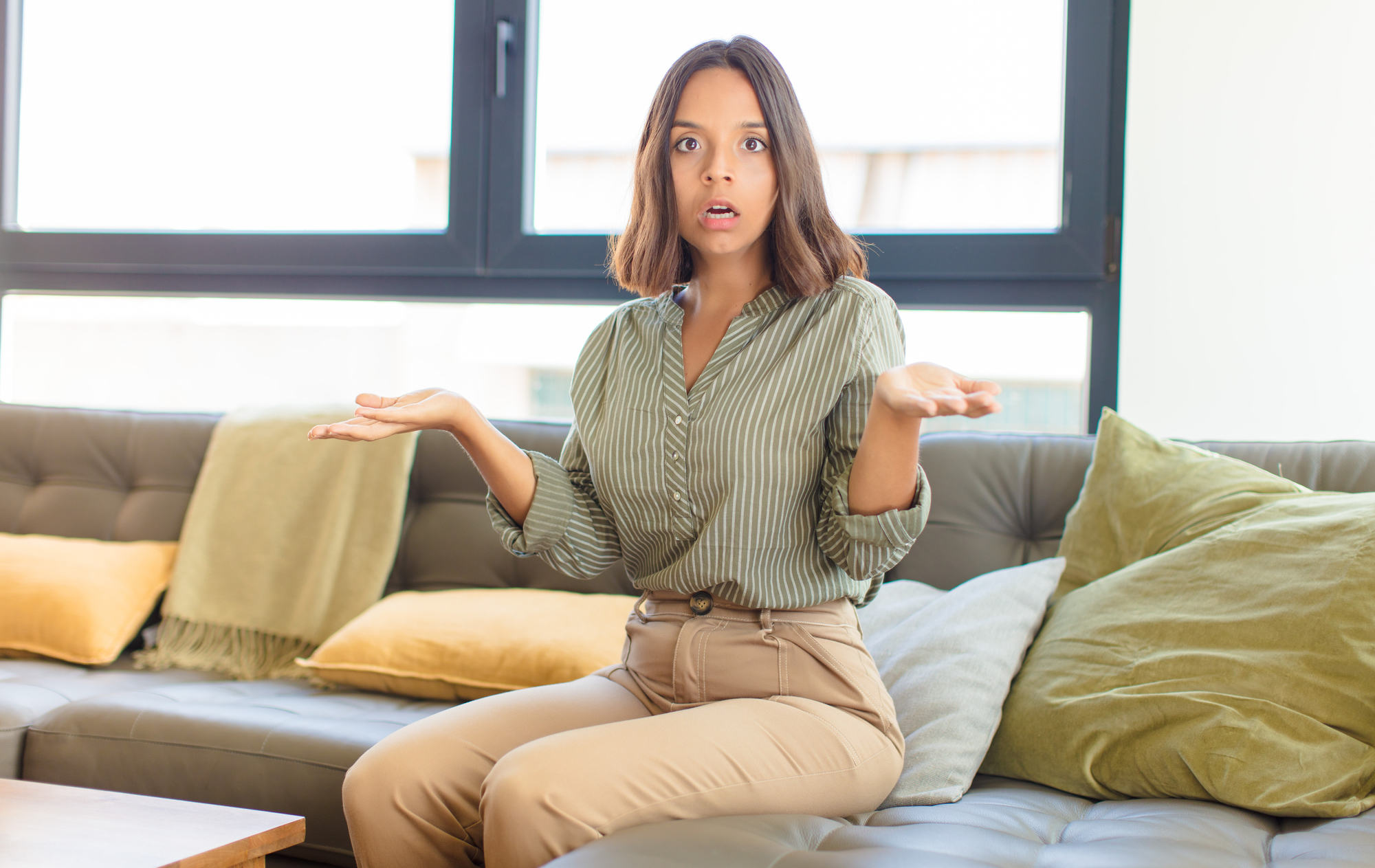 A woman with shoulder-length brown hair and a green striped blouse is sitting on a sofa with green and yellow cushions. She has an expression of surprise or confusion and is holding her hands out to the sides. There is a large window in the background.