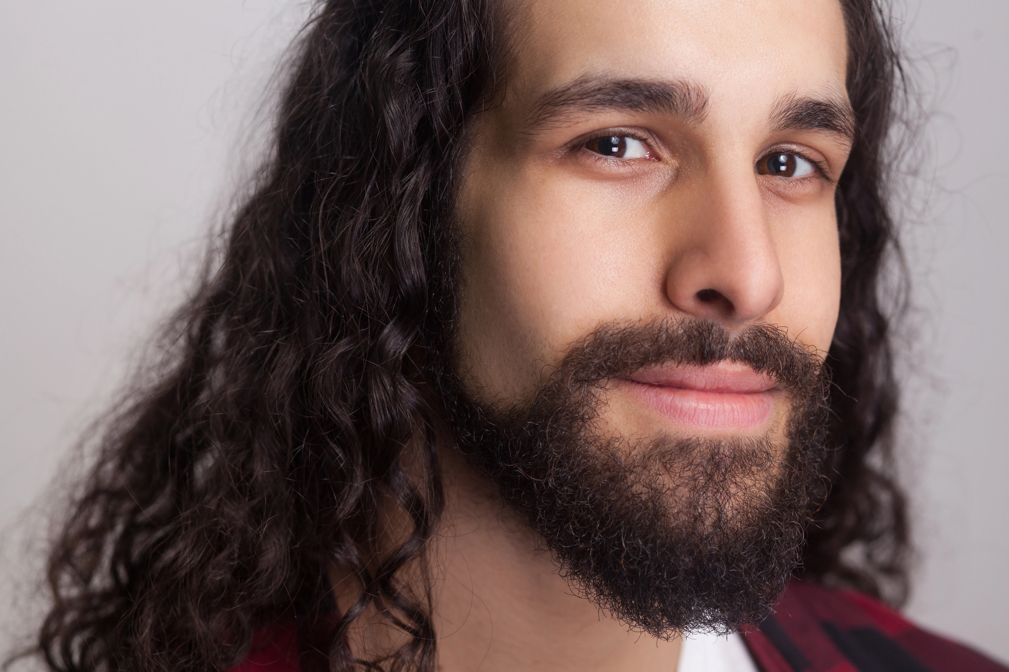 Close-up of a person with long, curly hair and a full beard, giving a gentle smile. The background is neutral and the lighting is soft, highlighting their facial features. The person is wearing a red and black checkered shirt.