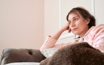 A woman with long hair, dressed in a light pink sweater, is sitting on a plush sofa, resting her head on her hand. She gazes thoughtfully into the distance. The room has soft, neutral tones and a cozy atmosphere.