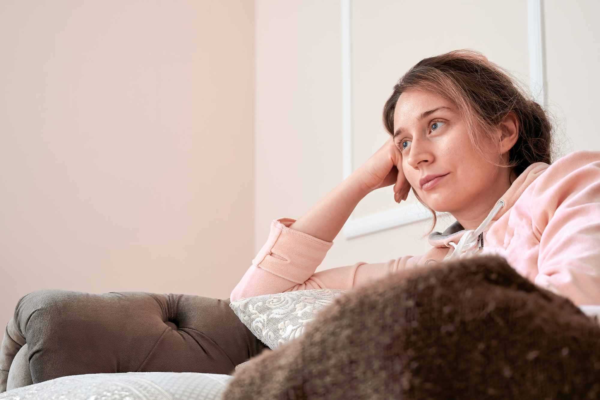 A woman with long hair, dressed in a light pink sweater, is sitting on a plush sofa, resting her head on her hand. She gazes thoughtfully into the distance. The room has soft, neutral tones and a cozy atmosphere.