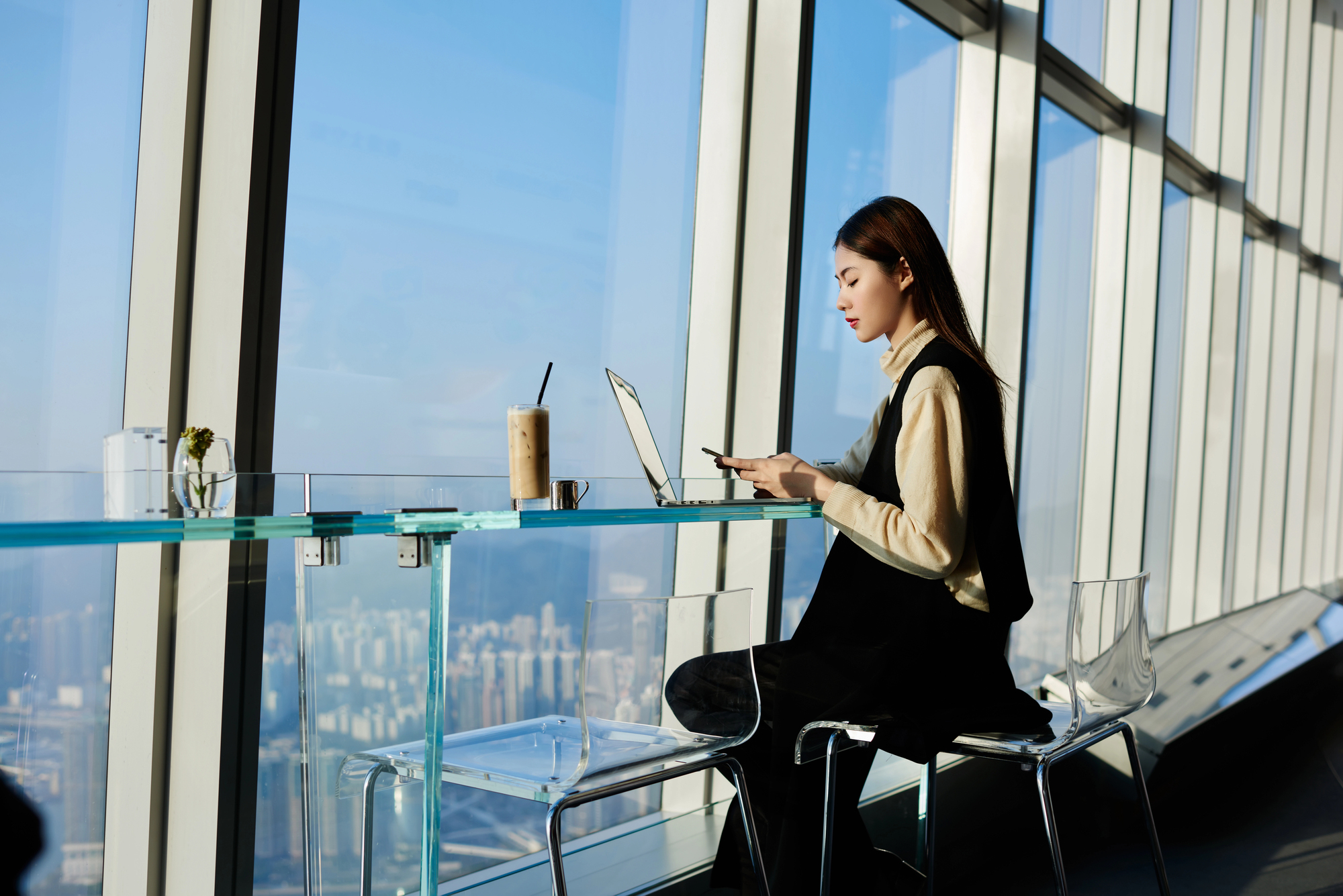 A woman sits at a high glass table near large windows, working on her laptop and holding a phone. Next to her is a tall iced coffee. The view outside the windows shows a city skyline under a clear blue sky. She is dressed in a light shirt and black dress.