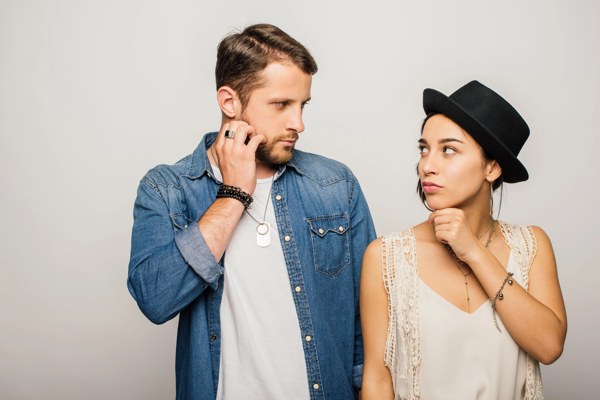 A man and woman stand against a neutral background, looking at each other with thoughtful expressions. The man is wearing a denim shirt and has his hand on his neck, while the woman, in a white top and black hat, has her hand under her chin.