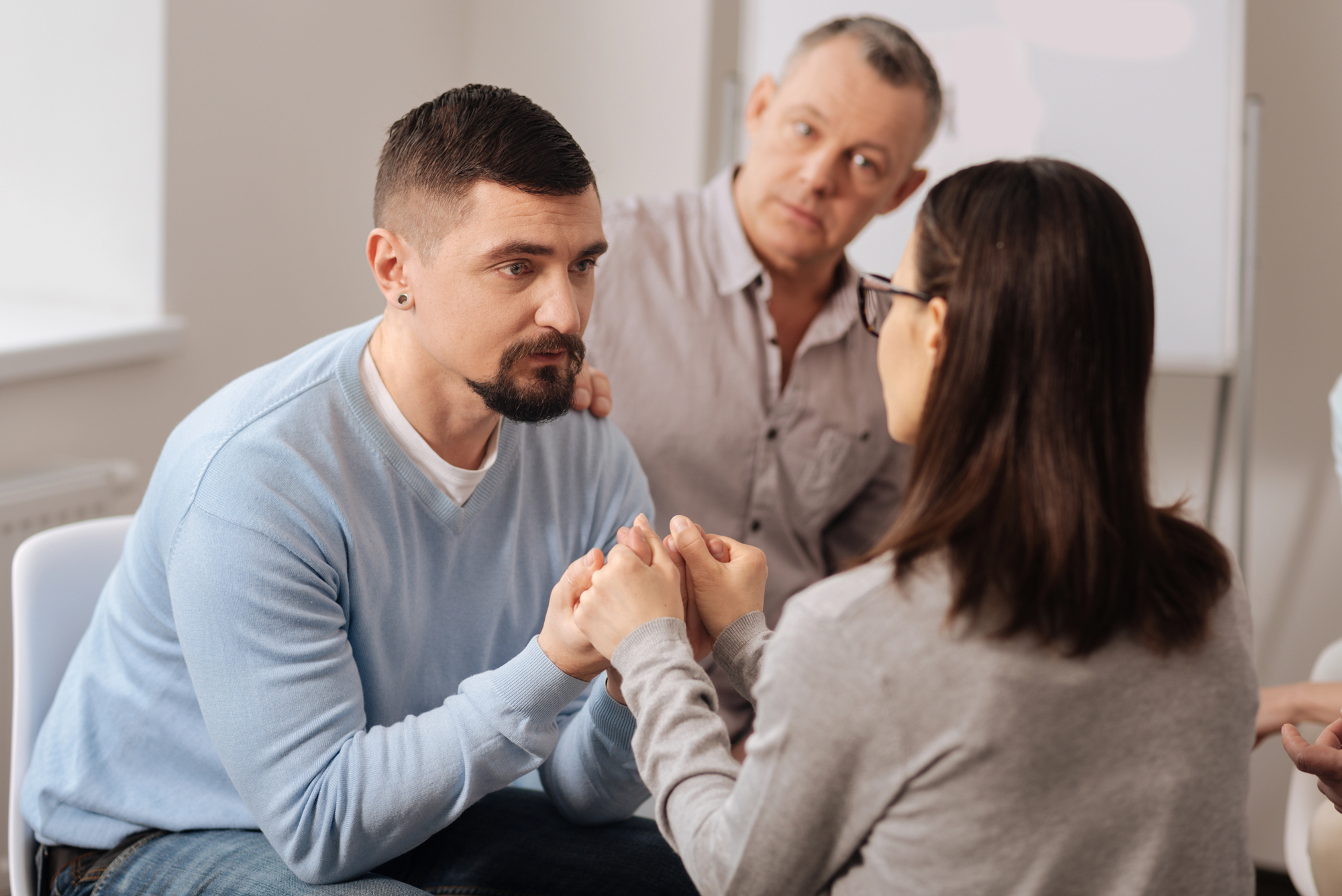 Three people are engaged in a serious conversation. A man with a trimmed beard wearing a light blue sweater holds hands with a woman in a gray sweater and glasses, making eye contact. Another man in a gray shirt is sitting in the background, attentively watching.