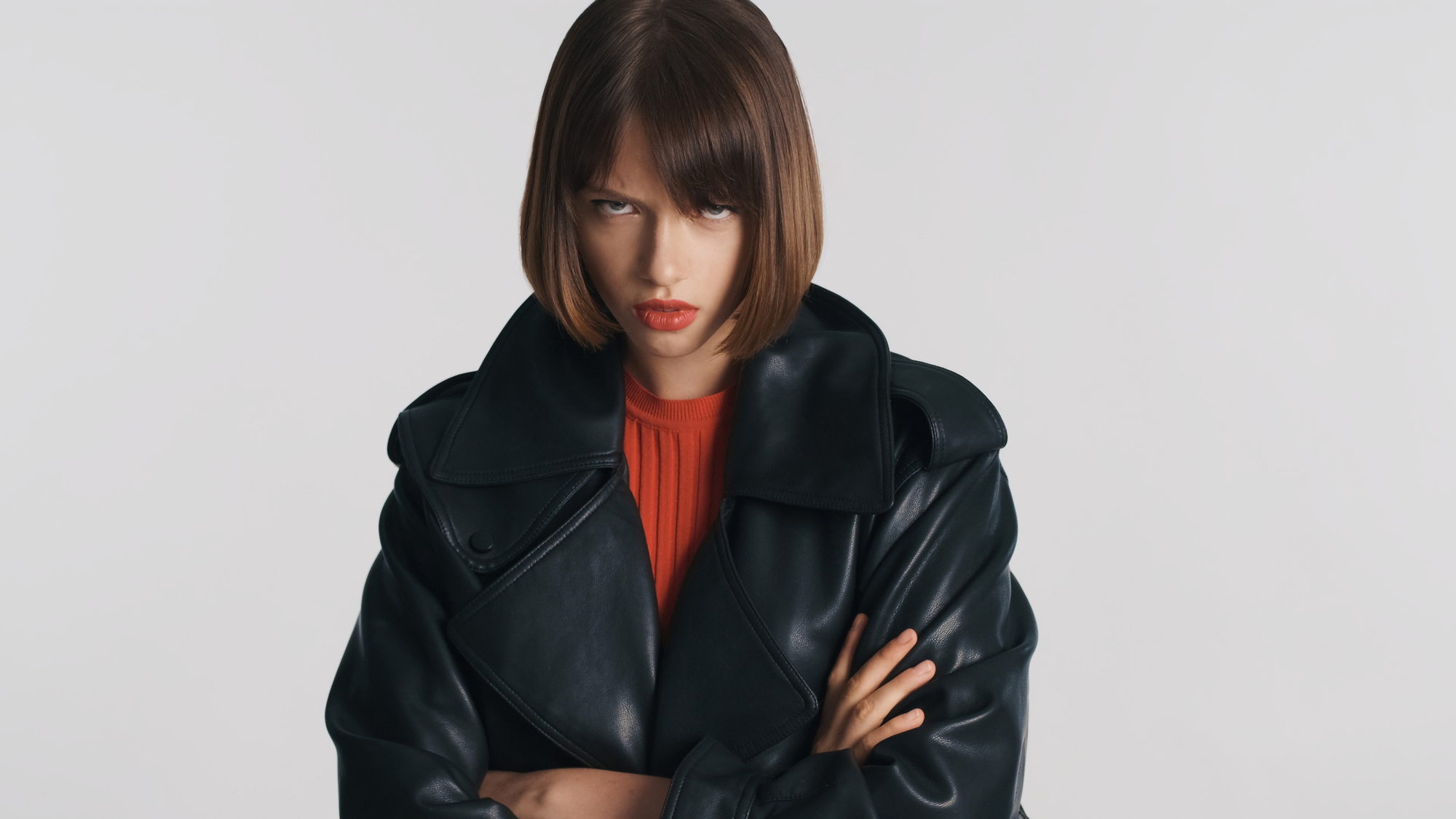 A person with shoulder-length brown hair and red lipstick is wearing a black leather jacket over a red shirt. They are standing against a plain white background with their arms crossed and a serious expression on their face.