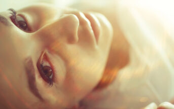 Close-up of a person's face lying down, illuminated by soft, warm light. The image focuses on one eye and part of the nose and mouth, with an ethereal and dreamy atmosphere created by light streaks and a shallow depth of field.