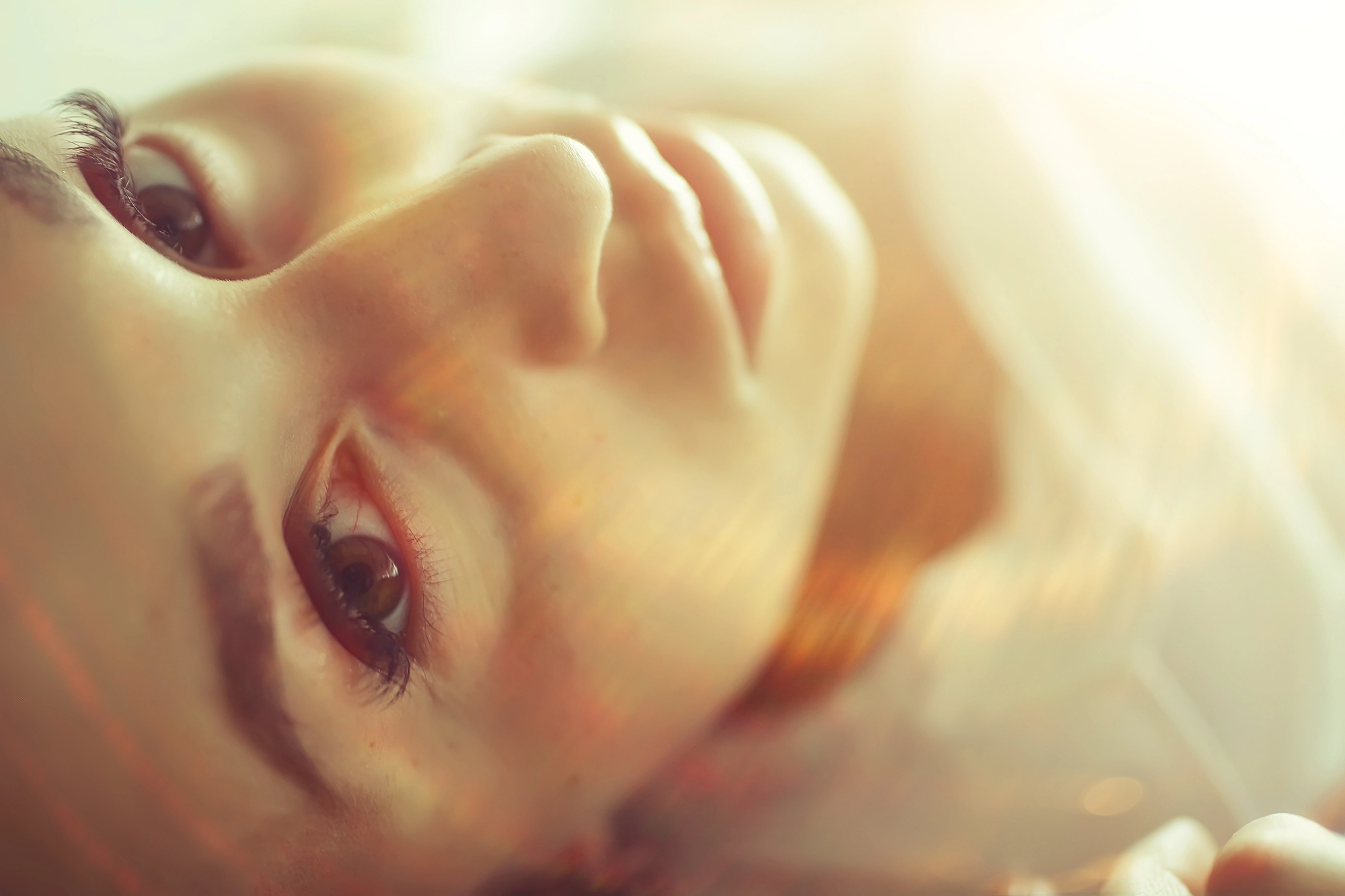 Close-up of a person's face lying down, illuminated by soft, warm light. The image focuses on one eye and part of the nose and mouth, with an ethereal and dreamy atmosphere created by light streaks and a shallow depth of field.