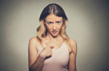 A woman with long, wavy blonde hair is looking directly at the camera with a surprised and slightly puzzled expression. She is wearing a light pink tank top and pointing at herself with her right hand. The background is a plain grey wall.