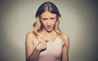 A woman with long, wavy blonde hair is looking directly at the camera with a surprised and slightly puzzled expression. She is wearing a light pink tank top and pointing at herself with her right hand. The background is a plain grey wall.