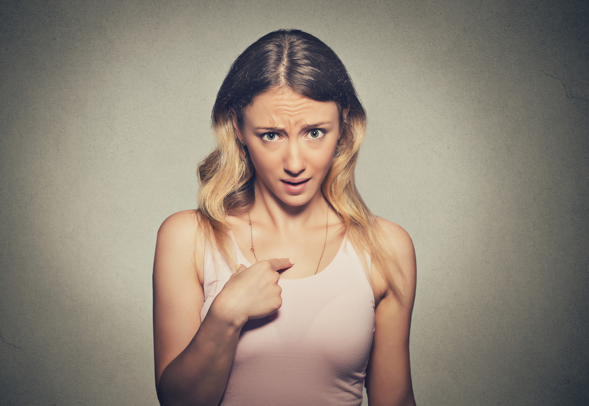 A woman with long, wavy blonde hair is looking directly at the camera with a surprised and slightly puzzled expression. She is wearing a light pink tank top and pointing at herself with her right hand. The background is a plain grey wall.