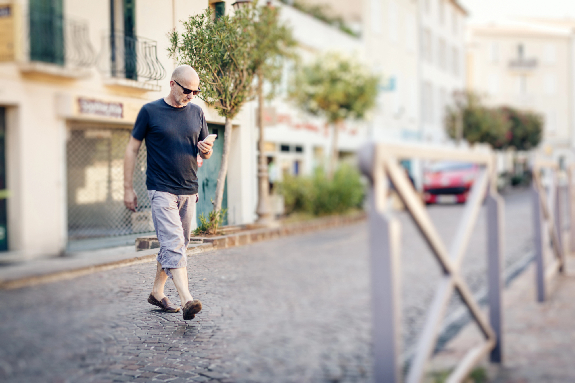 A bald man wearing sunglasses, a navy blue t-shirt, light grey shorts, and brown shoes is walking on a cobblestone street while looking at his smartphone. The background features buildings, trees, and a blurred-out red car.