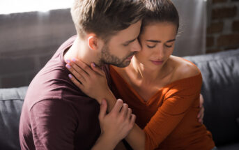 A man and a woman are sitting closely on a couch in a cozy environment. The man has his arm around the woman, who looks upset with her eyes closed and hand on her neck. The man appears to be comforting her, with his eyes closed and forehead touching hers.