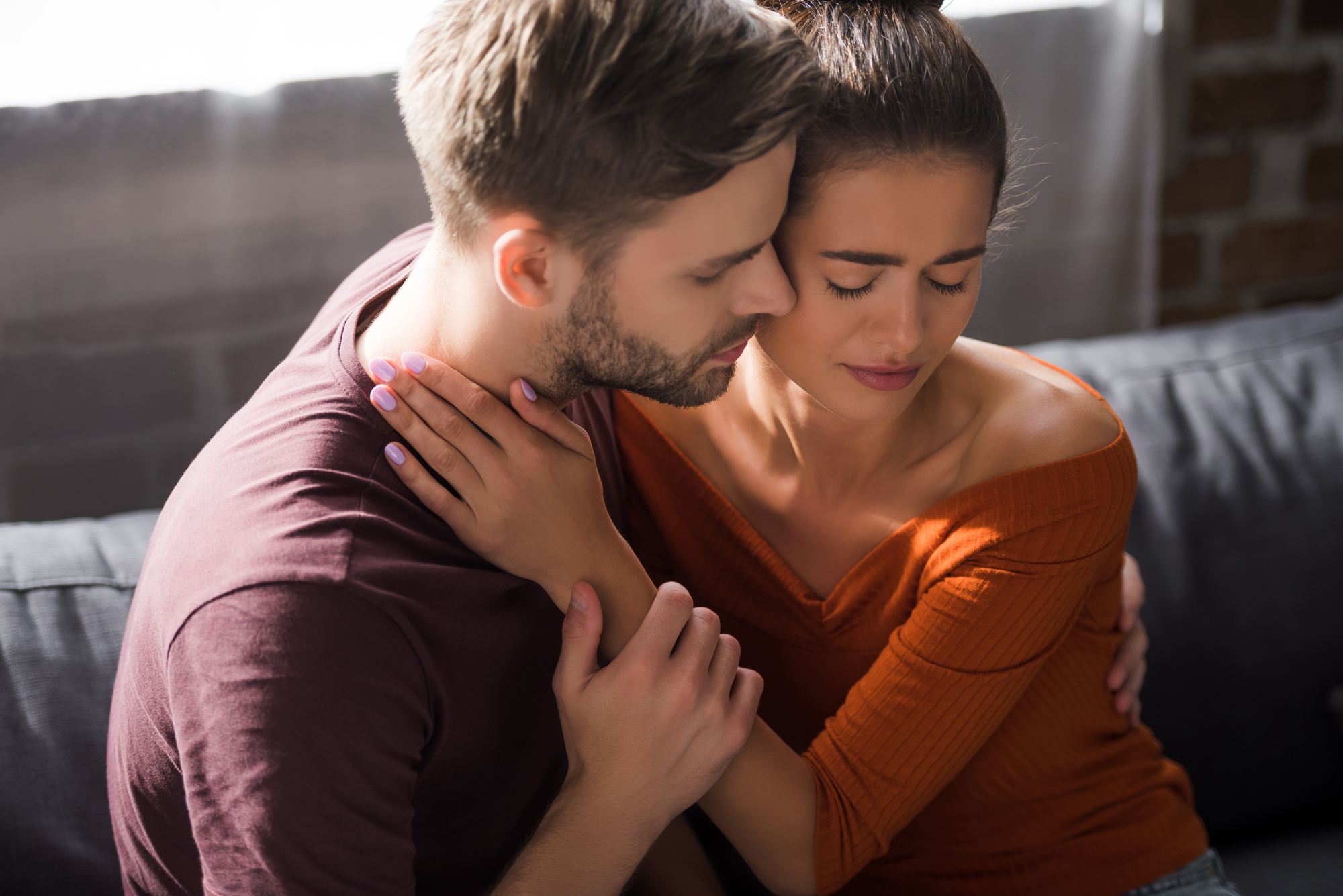 A man and a woman are sitting closely on a couch in a cozy environment. The man has his arm around the woman, who looks upset with her eyes closed and hand on her neck. The man appears to be comforting her, with his eyes closed and forehead touching hers.