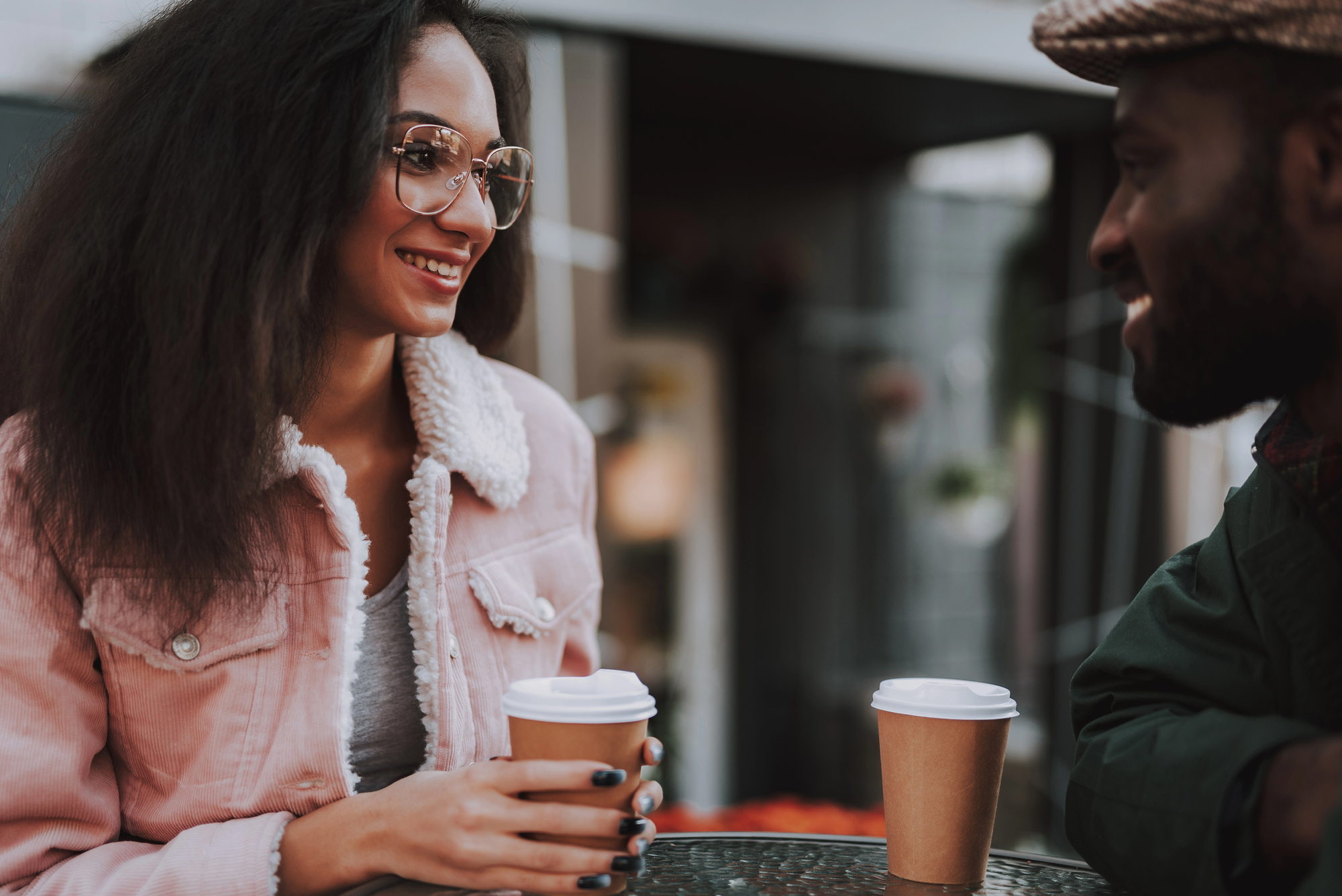 A woman with wavy hair and glasses, wearing a pink jacket, smiles while holding a to-go coffee cup and sitting at an outdoor café table across from a man in a green jacket and brown hat, who also has a to-go coffee cup.