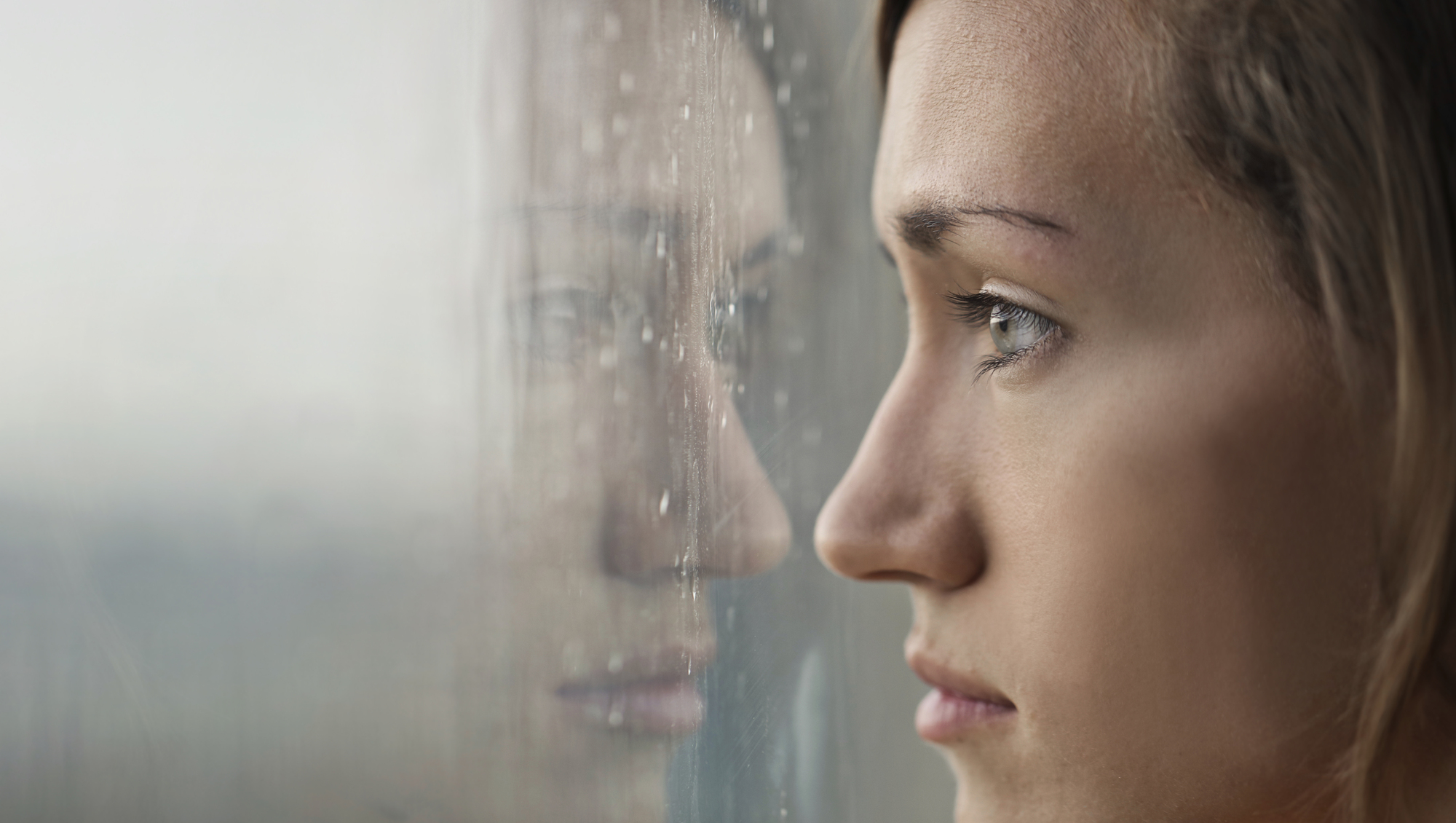 A close-up of a person's face looking out a window on a rainy day. The window is wet with rain droplets, and the person's reflection is visible in the glass, showing a contemplative and somber expression.