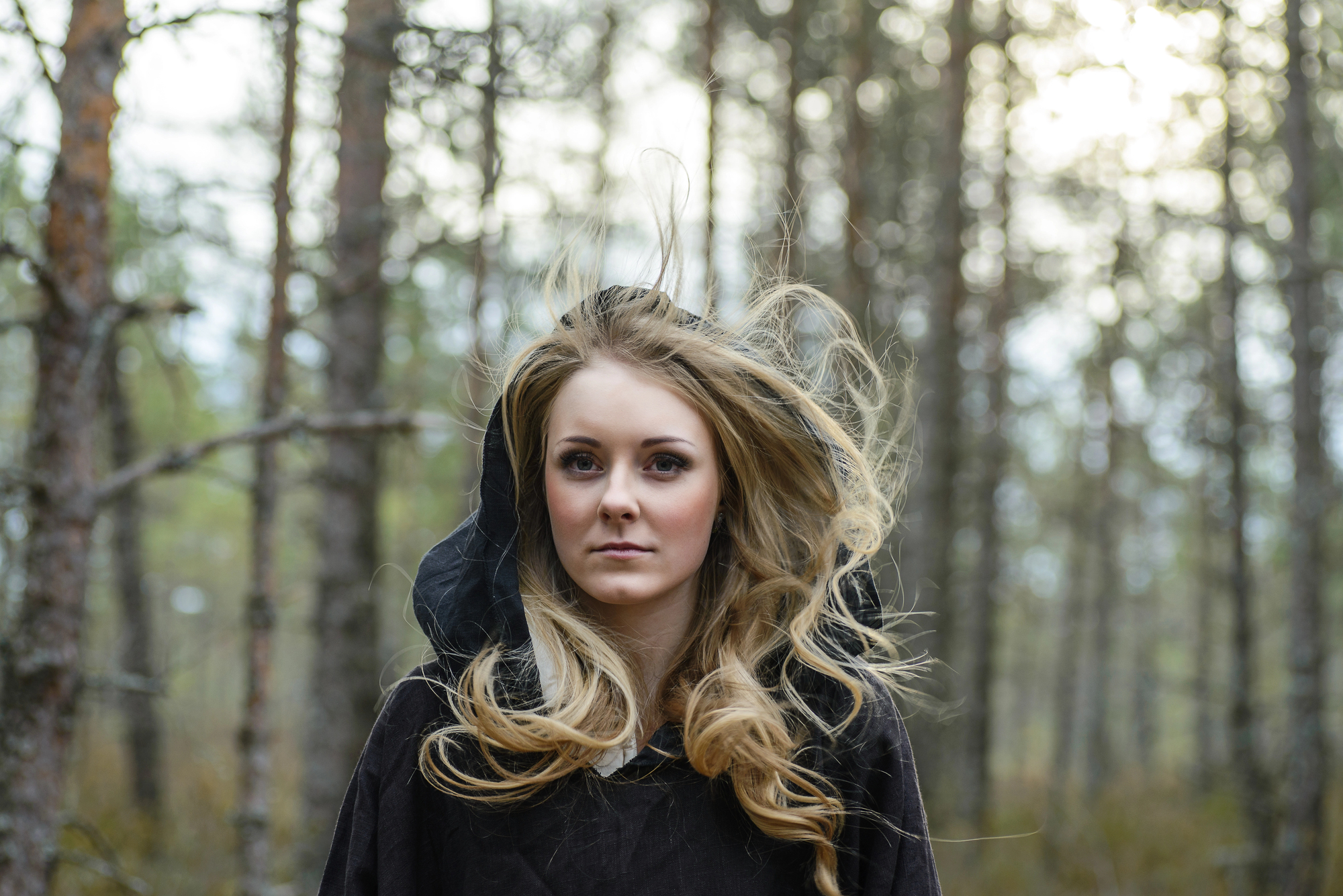 A woman with long, wavy blonde hair wearing a dark hooded cloak stands in a forest. Her hair is windswept, and the background shows blurry trees with a hint of light filtering through. Her expression is calm and focused.