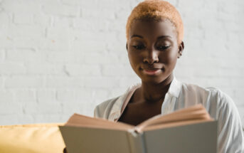 A person with short, light-colored hair is seated and reading a book against a light brick wall. They are wearing a white shirt and appear to be focused on the book.