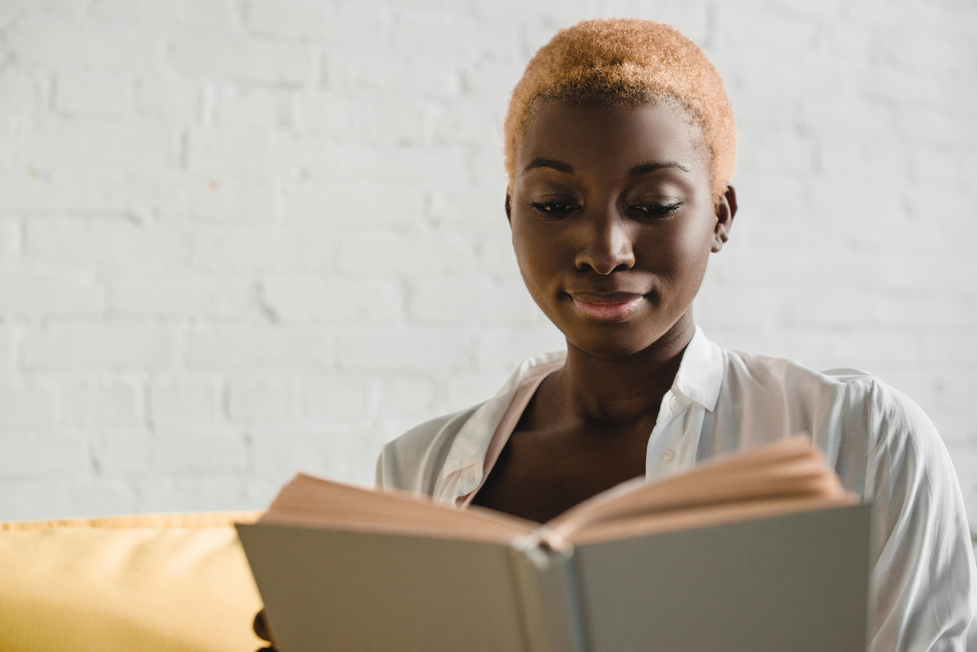 A person with short, light-colored hair is seated and reading a book against a light brick wall. They are wearing a white shirt and appear to be focused on the book.