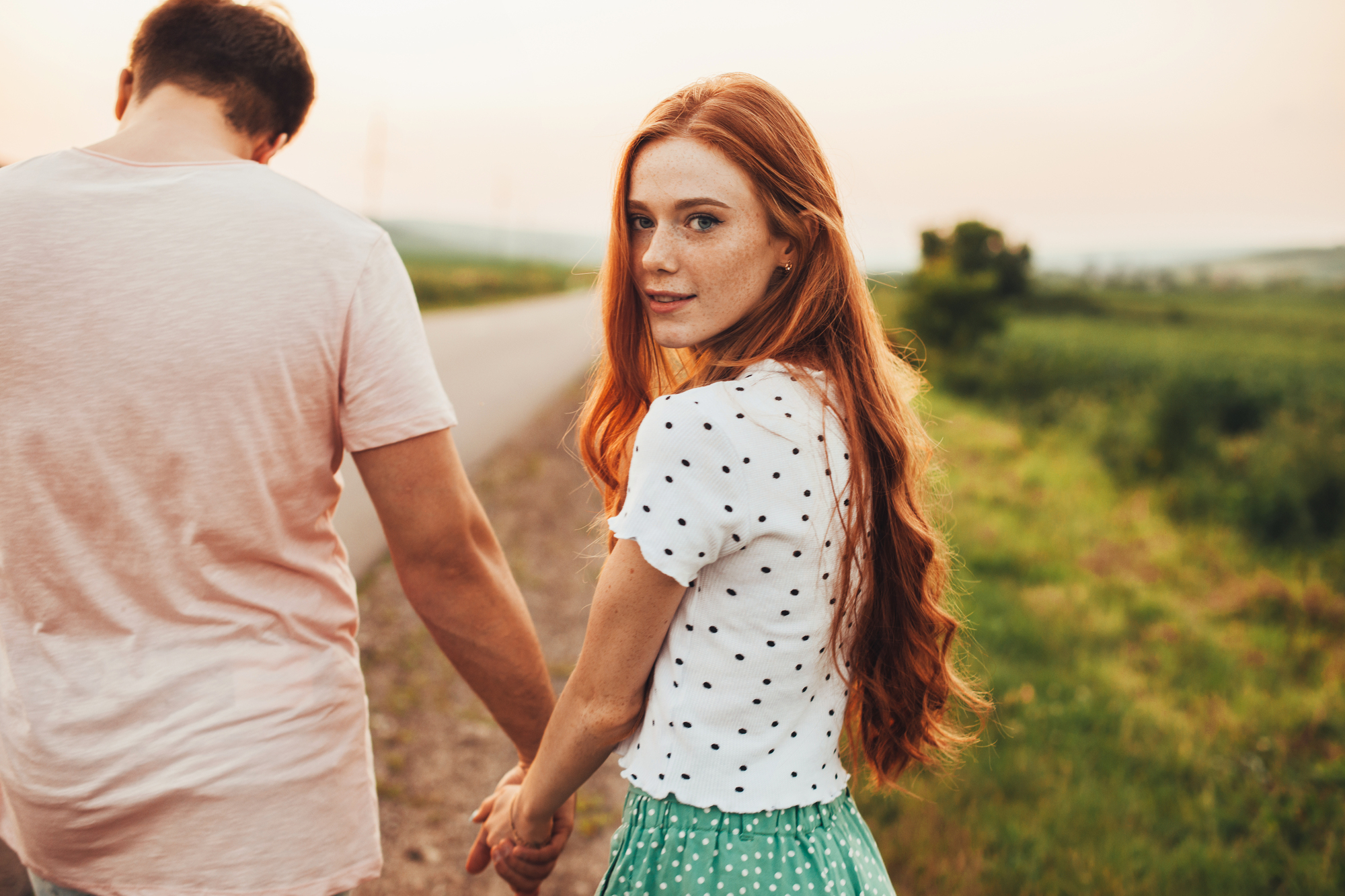 A young woman with long red hair and wearing a white polka dot shirt and green skirt holds hands with a man in a light pink shirt while walking on a path through a grassy field at sunset. She looks over her shoulder towards the camera.