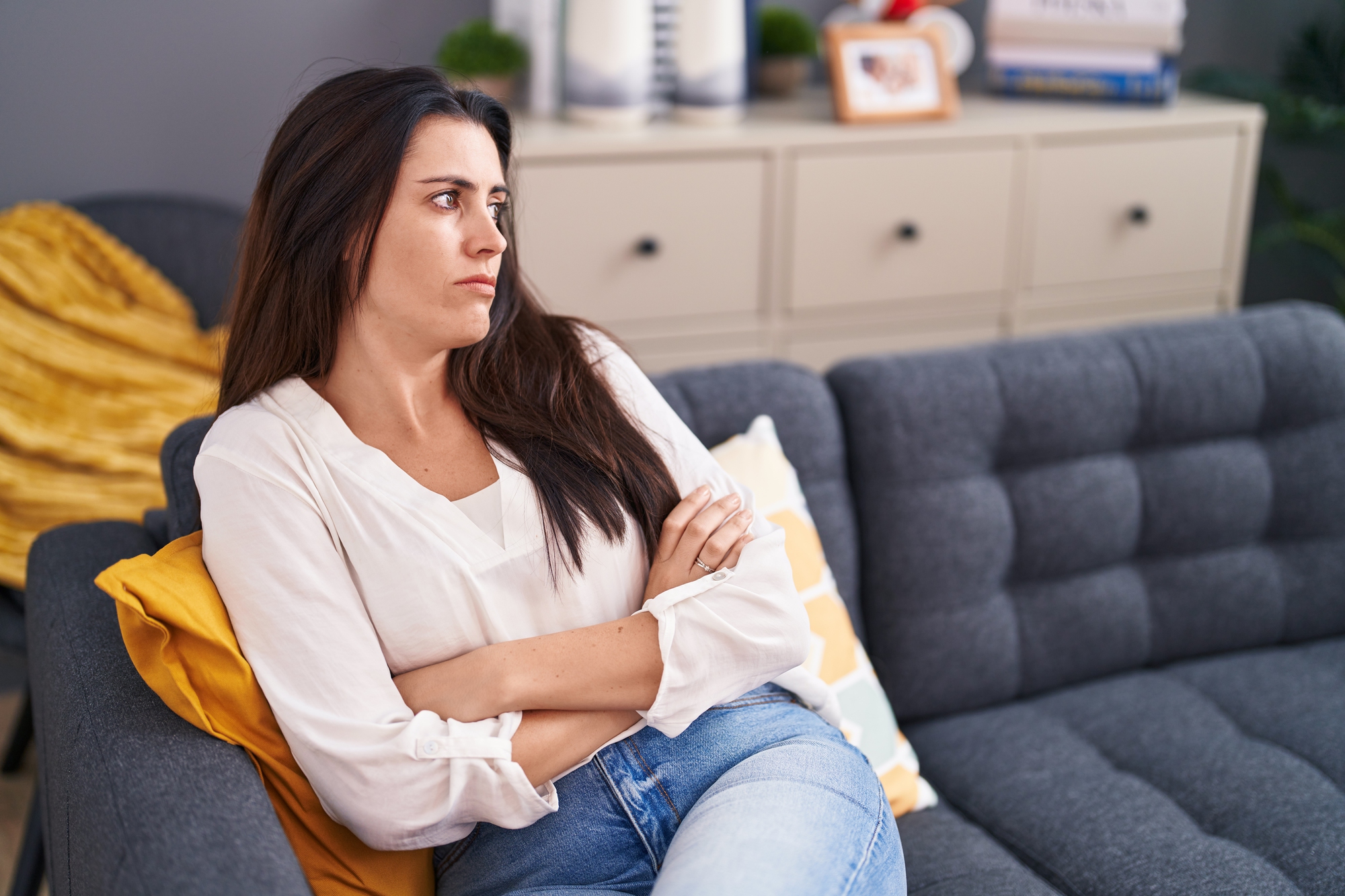 A woman with long dark hair is sitting on a gray couch, looking to the side with a pensive expression. She is wearing a white blouse and blue jeans, with her arms crossed. There is yellow blanket beside her and a wooden cabinet with decor in the background.