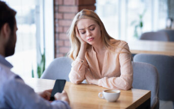 A woman with long blonde hair wearing a light pink blouse rests her head on her hand while sitting at a table in a cafe, looking tired or bored. In the foreground, a man is sitting across from her, focusing on his phone. A cup of coffee is on the table.