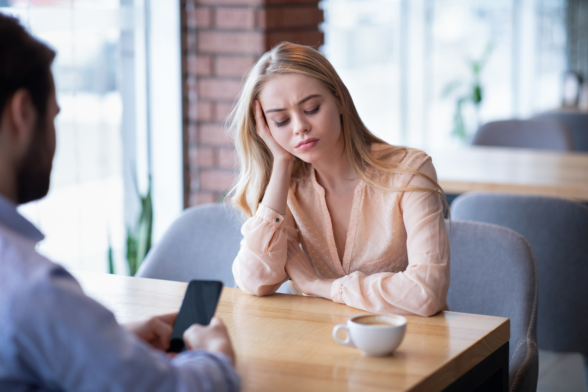 A woman with long blonde hair wearing a light pink blouse rests her head on her hand while sitting at a table in a cafe, looking tired or bored. In the foreground, a man is sitting across from her, focusing on his phone. A cup of coffee is on the table.