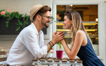 A smiling couple holding hands across a café table, gazing into each other's eyes. The man, wearing a hat and glasses, sits opposite the woman with sunglasses on her head. A small potted plant is on the table outside a cozy establishment with colorful flowers in the background.