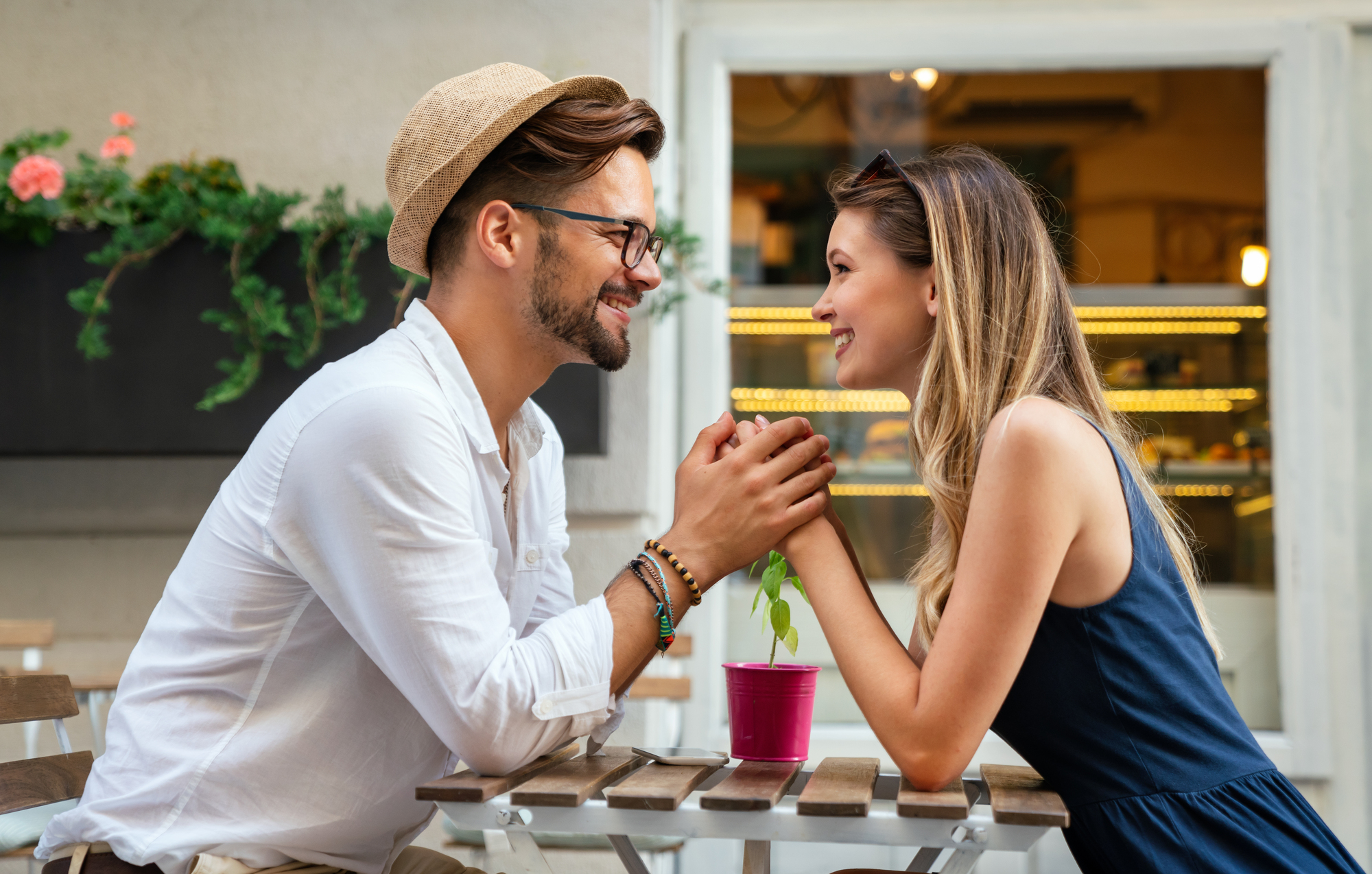 A smiling couple holding hands across a café table, gazing into each other's eyes. The man, wearing a hat and glasses, sits opposite the woman with sunglasses on her head. A small potted plant is on the table outside a cozy establishment with colorful flowers in the background.