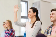 A young woman in a classroom raising her hand, smiling. She has brown hair tied back and is wearing a pink and gray top. Other students are seated around her, some smiling and looking attentive. The room is bright with natural light coming from windows in the background.