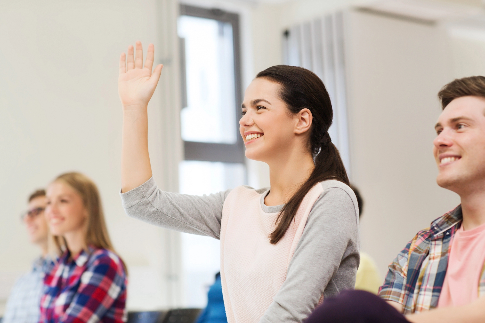 A young woman in a classroom raising her hand, smiling. She has brown hair tied back and is wearing a pink and gray top. Other students are seated around her, some smiling and looking attentive. The room is bright with natural light coming from windows in the background.