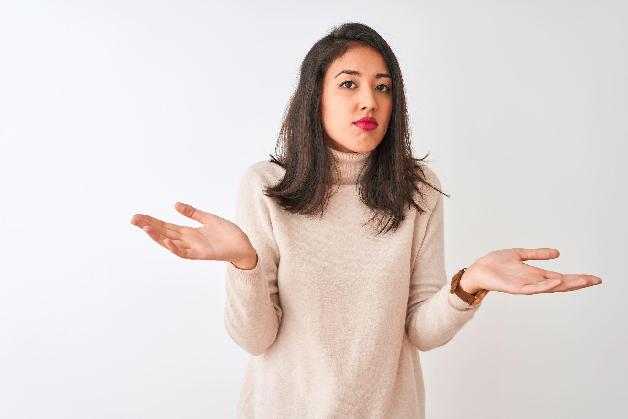 A young woman with shoulder-length dark hair, wearing a beige sweater, stands against a plain white background. She has a puzzled expression, with her eyebrows slightly raised and arms outstretched with palms up in a questioning gesture.