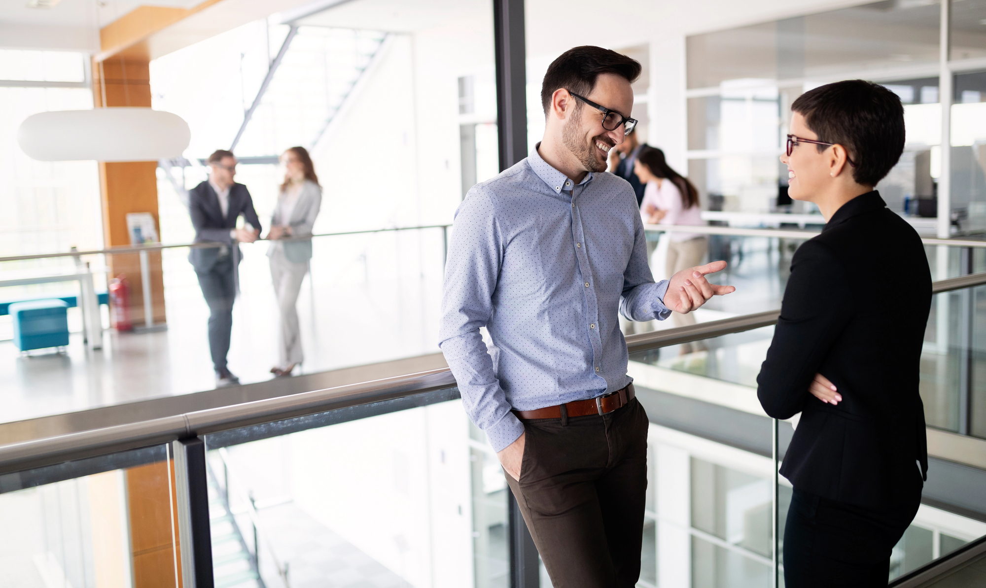 Two professionals in business attire have a friendly conversation on a bright and modern office balcony. In the background, two other pairs of colleagues are also engaged in discussions. The environment is open and airy, with contemporary decor.