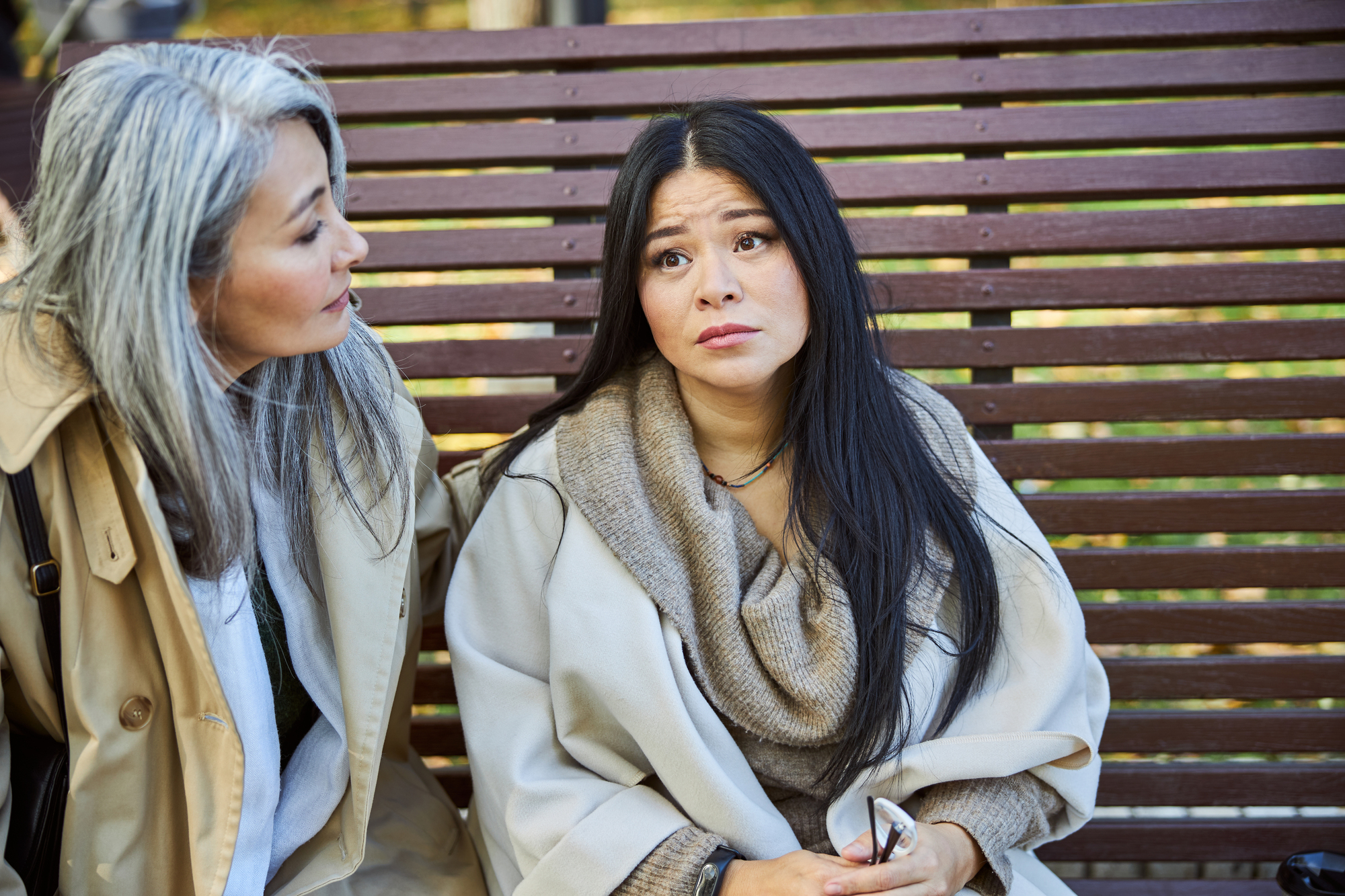 Two women are sitting together on a wooden bench. One woman with long, gray hair is talking to the other woman, who has long, black hair and is looking up with a thoughtful expression. Both are wearing beige coats. The background is slightly blurred but suggests an outdoor setting.