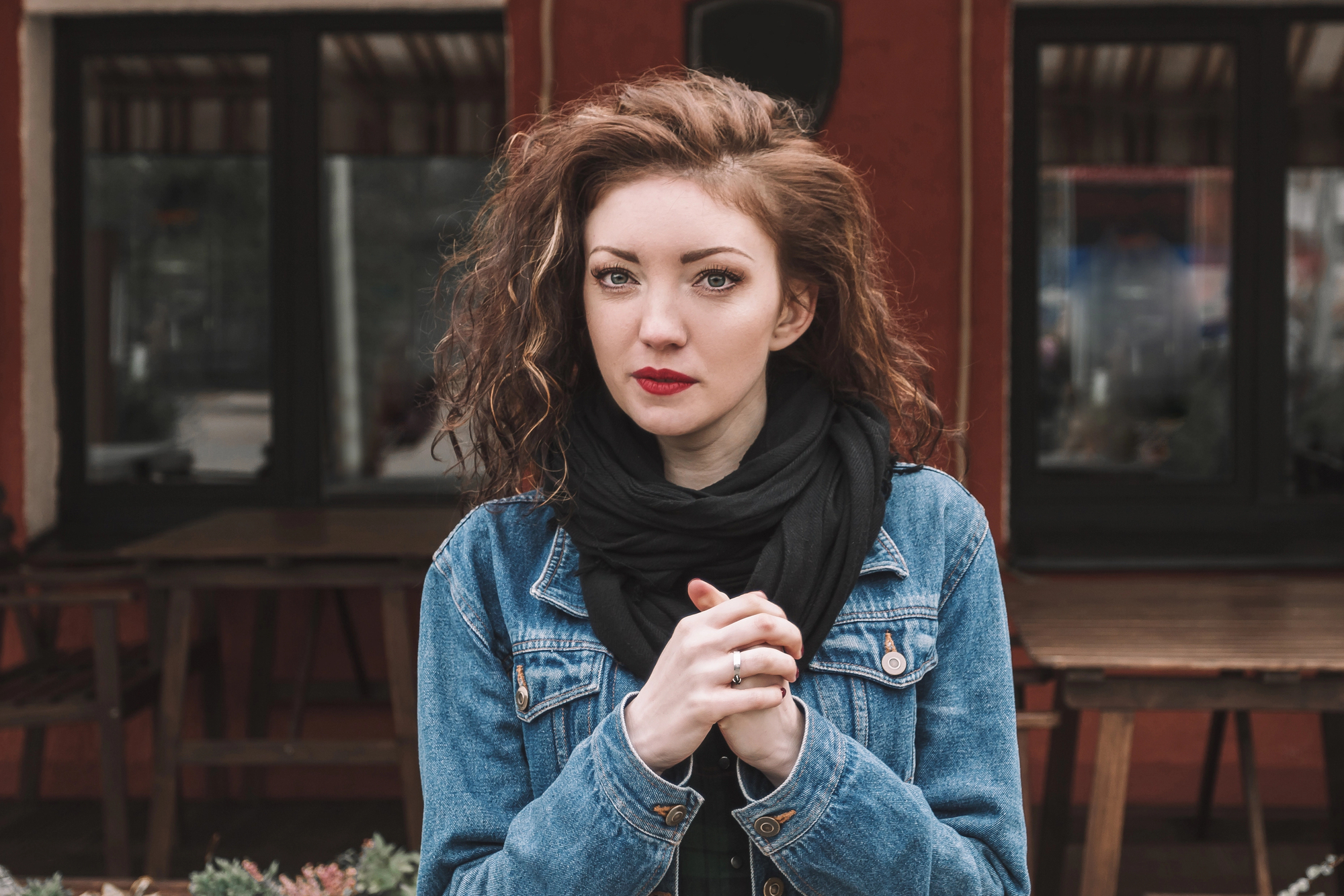 A woman with shoulder-length curly hair and red lipstick stands in front of a building with a red facade and dark-framed windows. She is wearing a denim jacket over a black scarf and appears to be clasping her hands together.