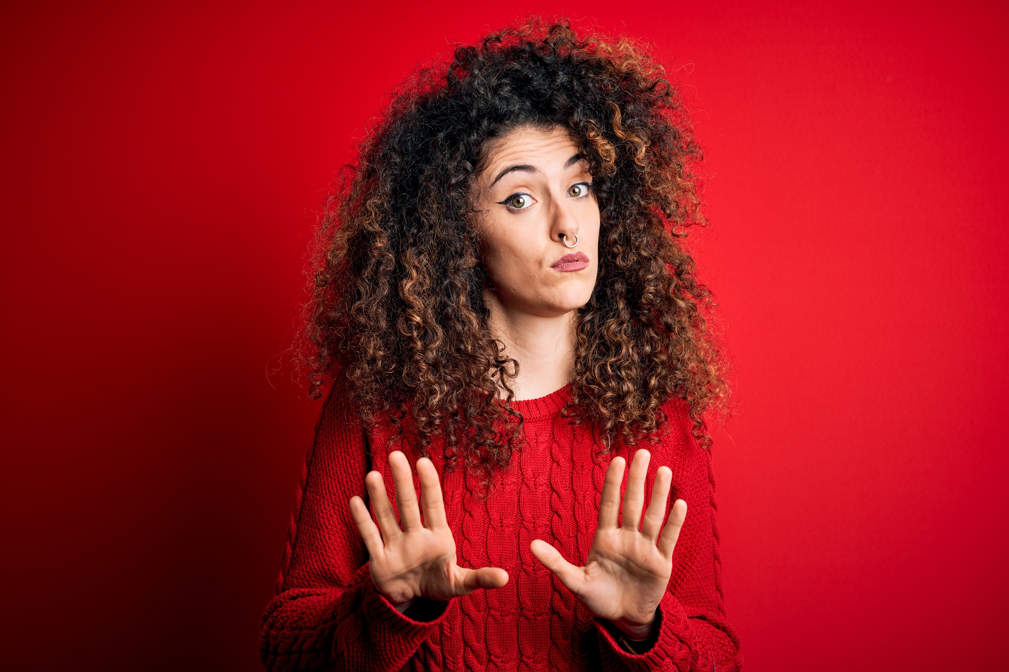 A woman with curly hair and a nose ring stands against a red background. She is wearing a red sweater and has a playful, slightly skeptical expression on her face. Both hands are raised with palms outward in a "stop" or "wait" gesture.