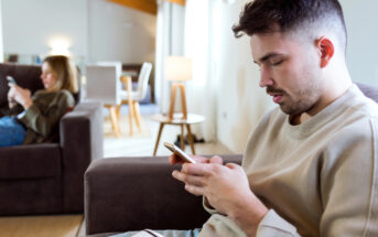 A man and woman sit in a living room, each absorbed in their smartphones. The man sits on a dark sofa in the foreground, wearing a light sweater, while the woman is on another sofa in the background, partially obscured by a table and lit lamp.