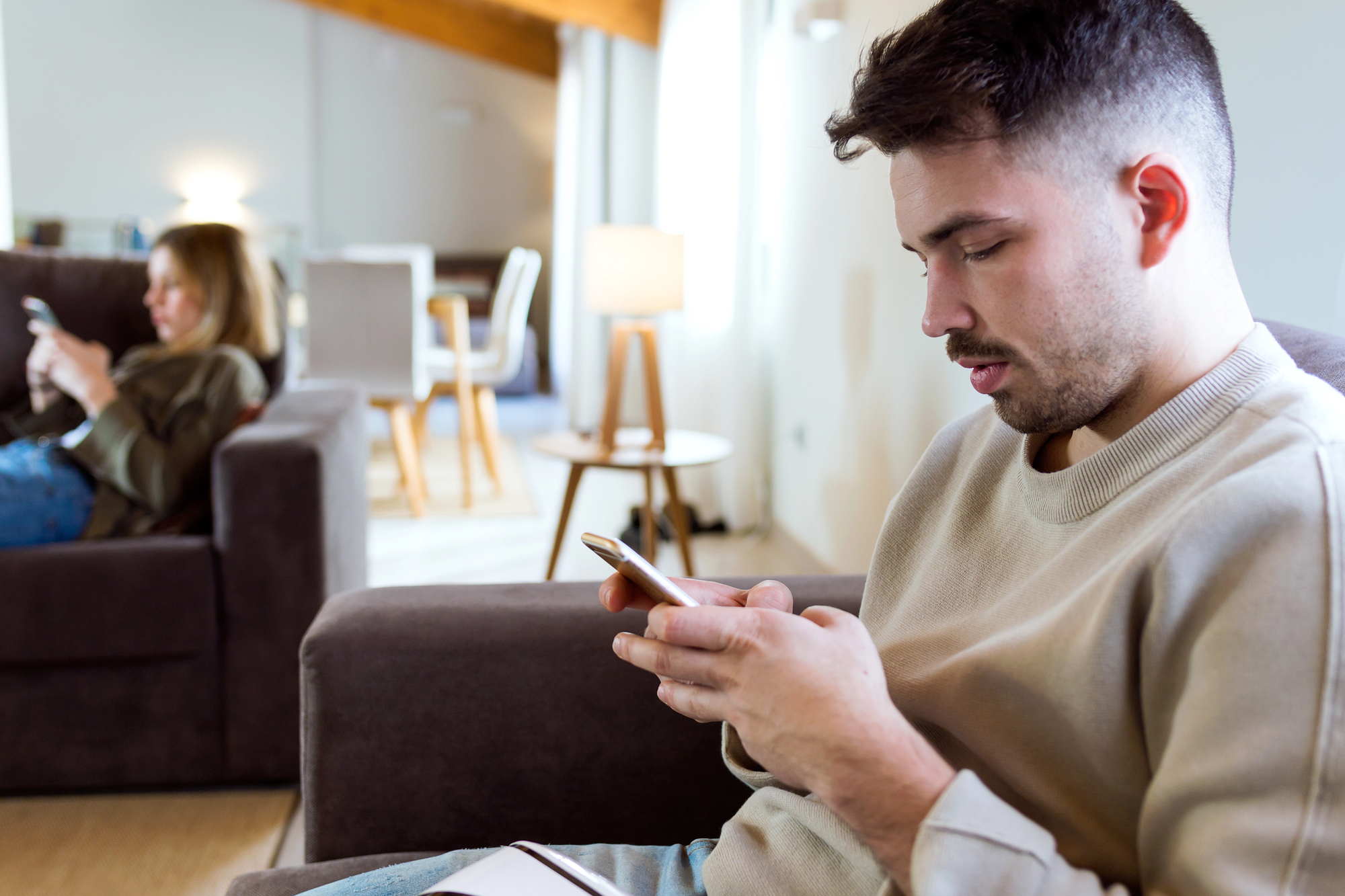 A man and woman sit in a living room, each absorbed in their smartphones. The man sits on a dark sofa in the foreground, wearing a light sweater, while the woman is on another sofa in the background, partially obscured by a table and lit lamp.