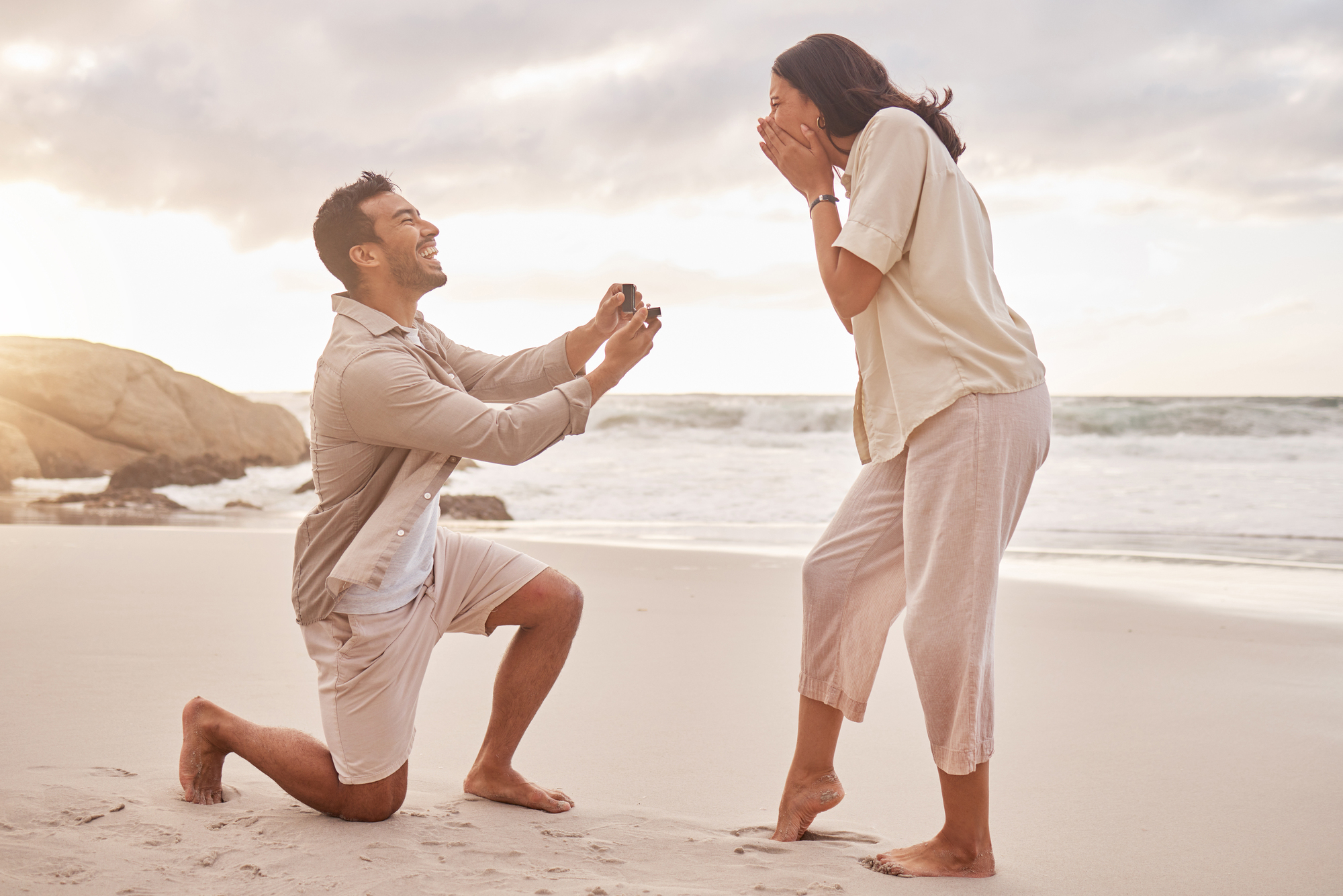 A man kneels on a sandy beach, proposing with a ring to a surprised woman who covers her mouth with her hands. The ocean and a cloudy sky are in the background, creating a romantic setting. Both are casually dressed in light-colored clothing.