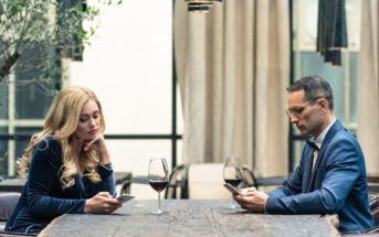 A woman with long blonde hair and a man wearing glasses sit across from each other at a wooden table, both looking at their smartphones. Two glasses of red wine are on the table. They appear to be in an outdoor or semi-outdoor setting with plants and curtains around.