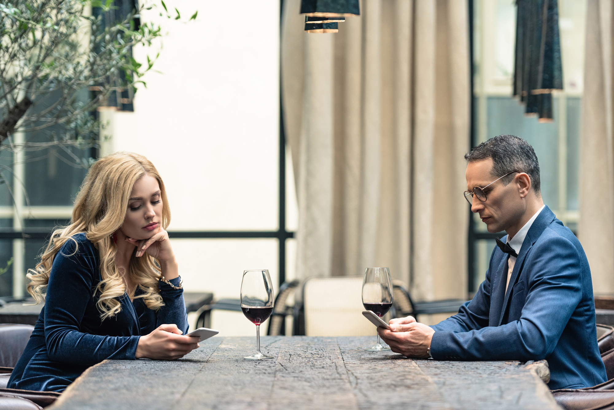 A woman with long blonde hair and a man wearing glasses sit across from each other at a wooden table, both looking at their smartphones. Two glasses of red wine are on the table. They appear to be in an outdoor or semi-outdoor setting with plants and curtains around.