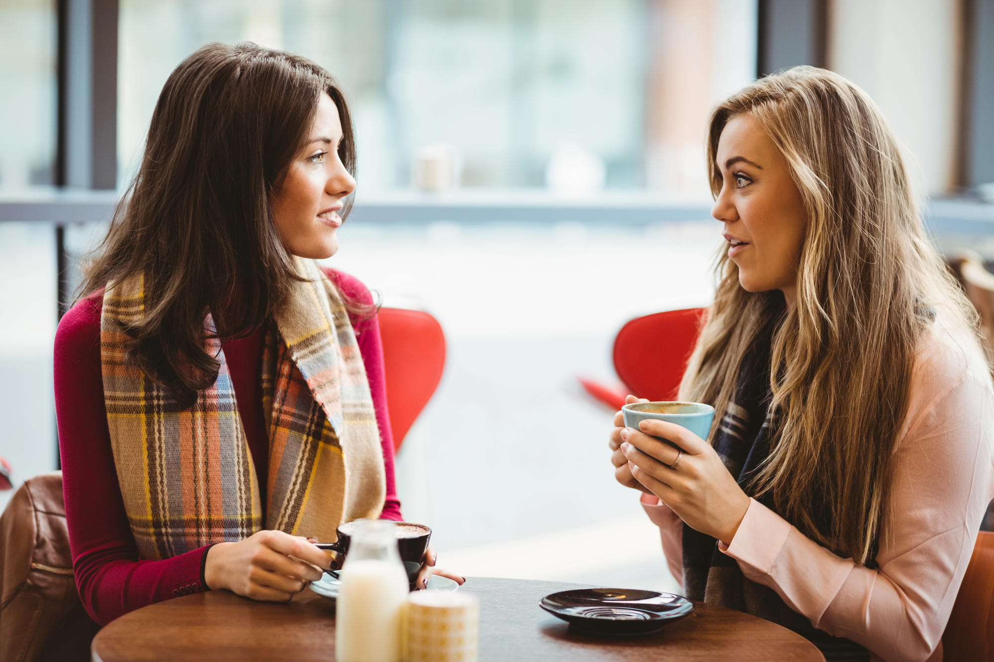 Two women are sitting at a cafe table, engaged in conversation. One woman is holding a blue coffee cup, while the other has a black cup on the table. Both are dressed warmly, with scarves. The background features a window with bright daylight outside.