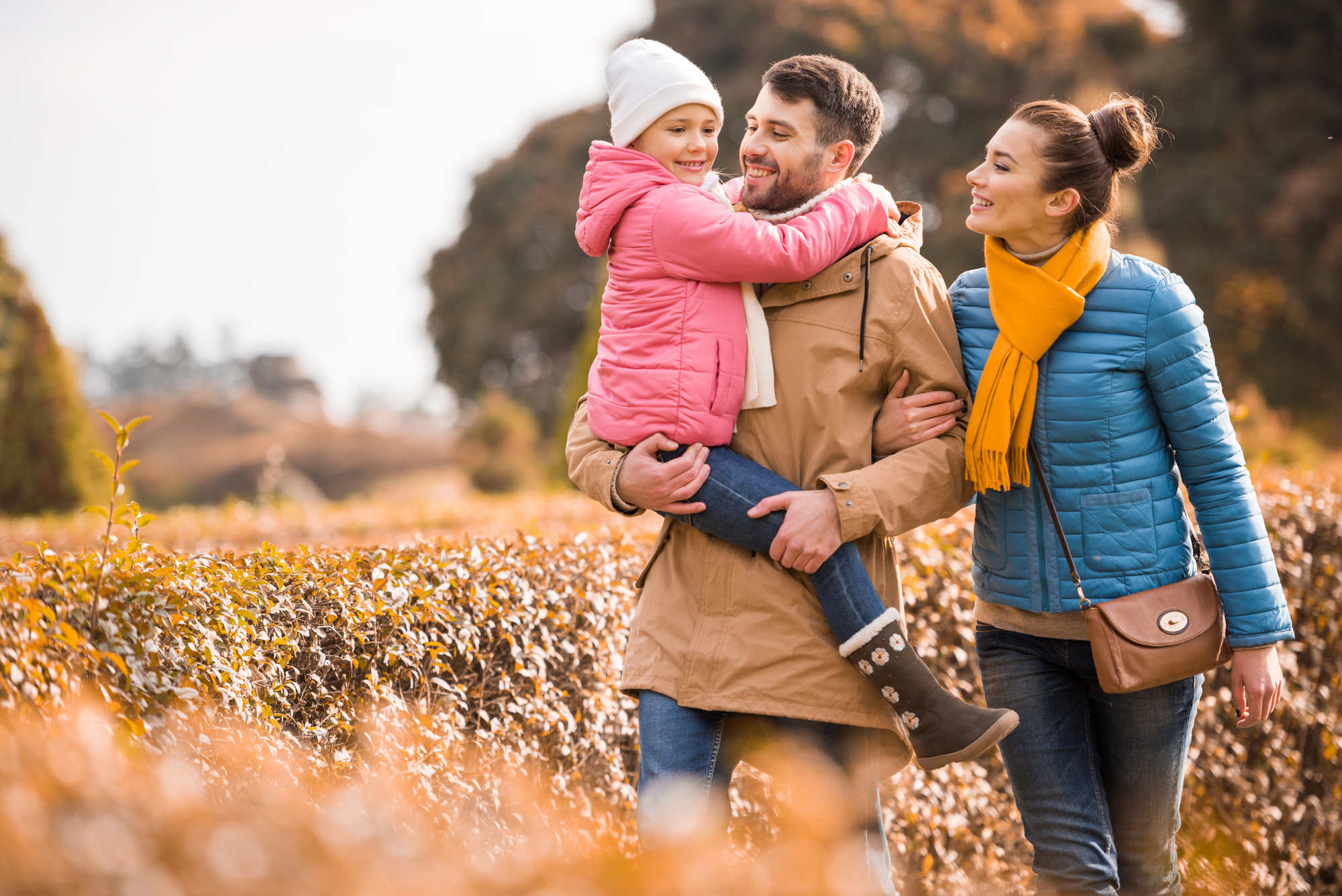 A family of three enjoying a walk in a park during autumn. A man in a tan coat carries a smiling child in a pink jacket and white beanie. A woman in a blue jacket and yellow scarf walks beside them, all surrounded by golden foliage.