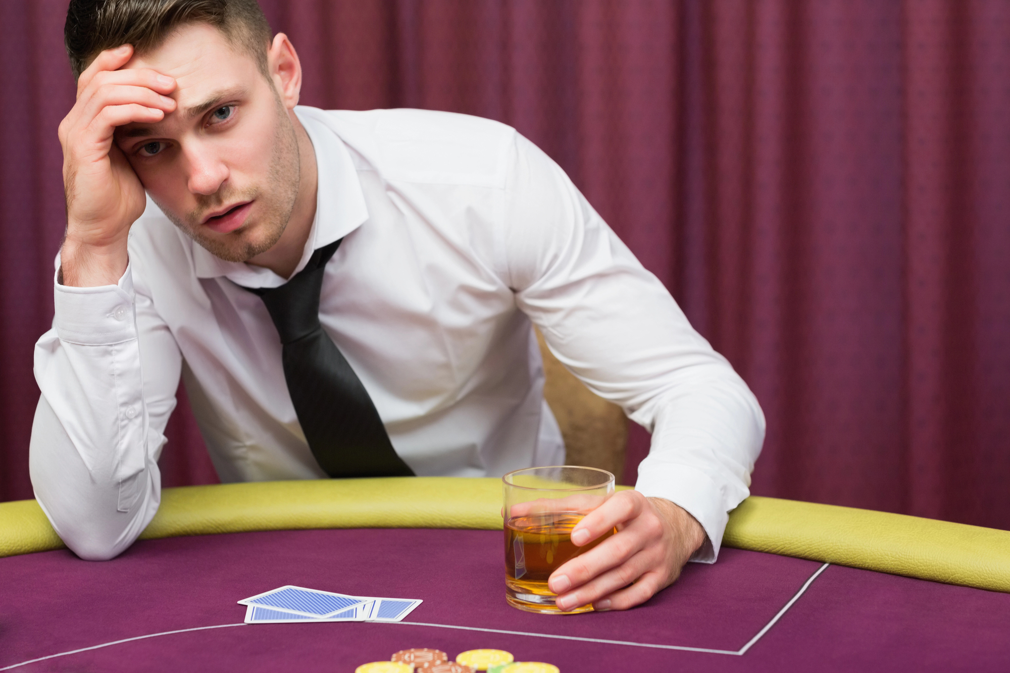 A man in a white dress shirt and black tie sits at a poker table, holding a drink in one hand and resting his head on the other. He appears distressed, with playing cards and poker chips spread on the table in front of him. A maroon curtain is in the background.