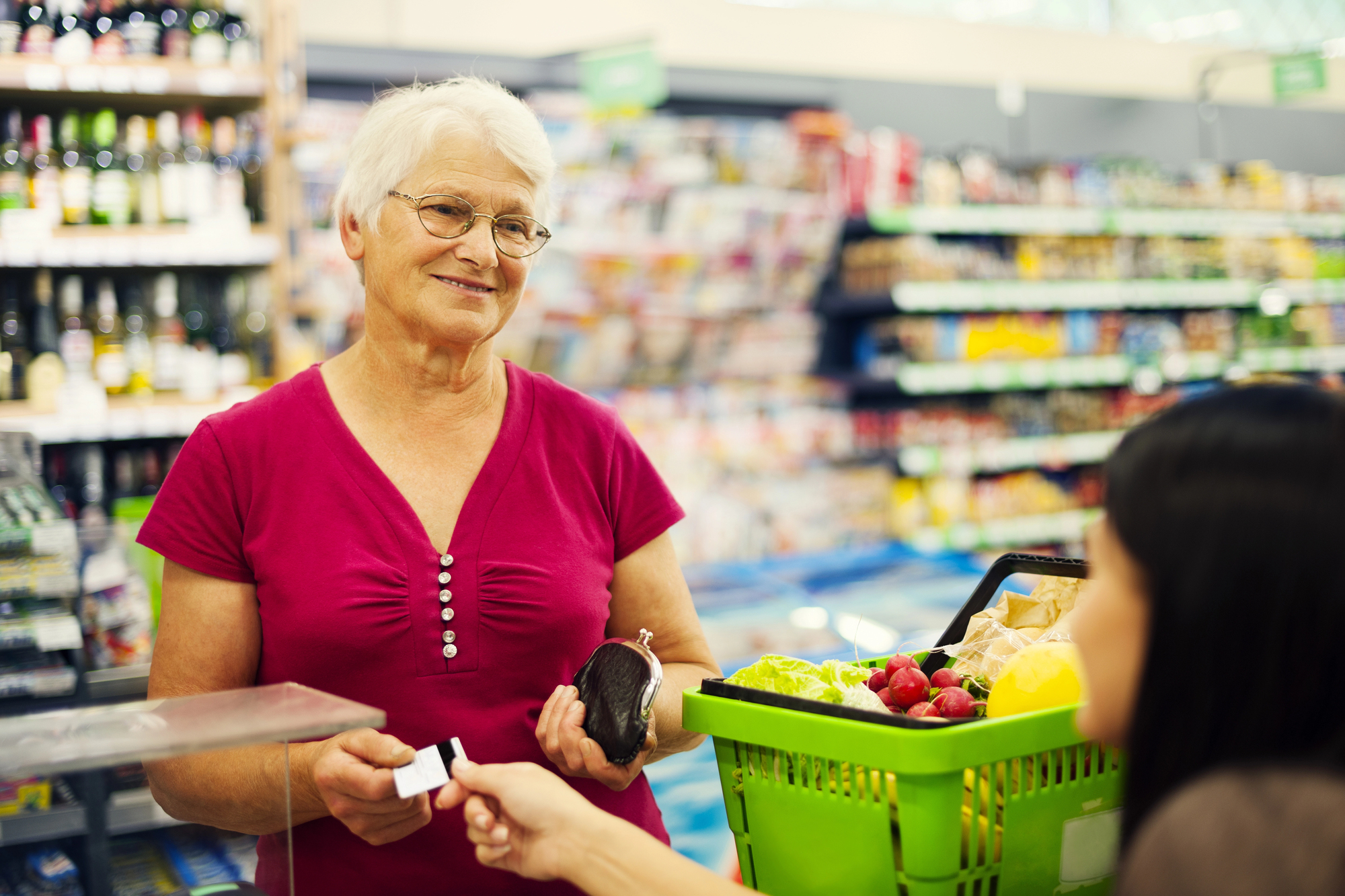 An elderly woman in a bright pink shirt is standing at a supermarket checkout counter, holding a wallet and an avocado. She is smiling and interacting with a person handing over a credit card. A green shopping basket with fruits and vegetables is on the counter.