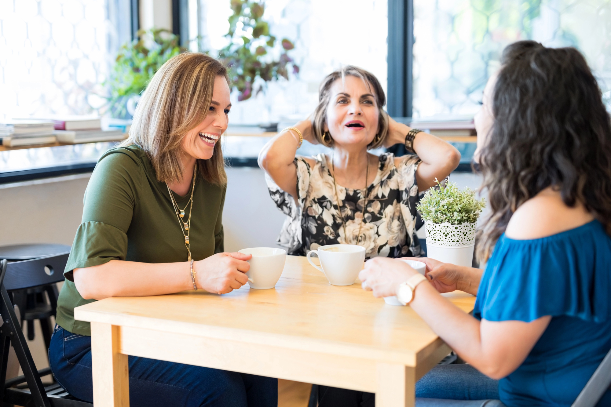 Three women are sitting at a table in a café, enjoying coffee and laughing together. The table has a small potted plant on it. The background shows large windows with sunlight streaming in and some greenery visible through the glass.