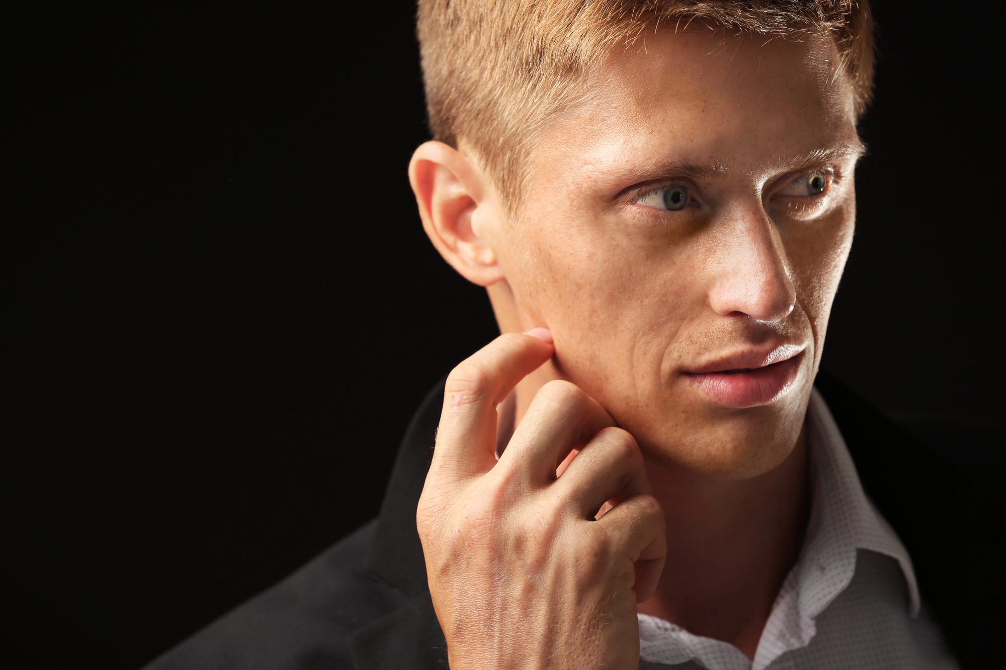 A man with short, light brown hair is gazing to the side. He is wearing a dark jacket and a white collared shirt. His hand is raised near his face, lightly touching his cheek. The background is dark, emphasizing his profile and thoughtful expression.