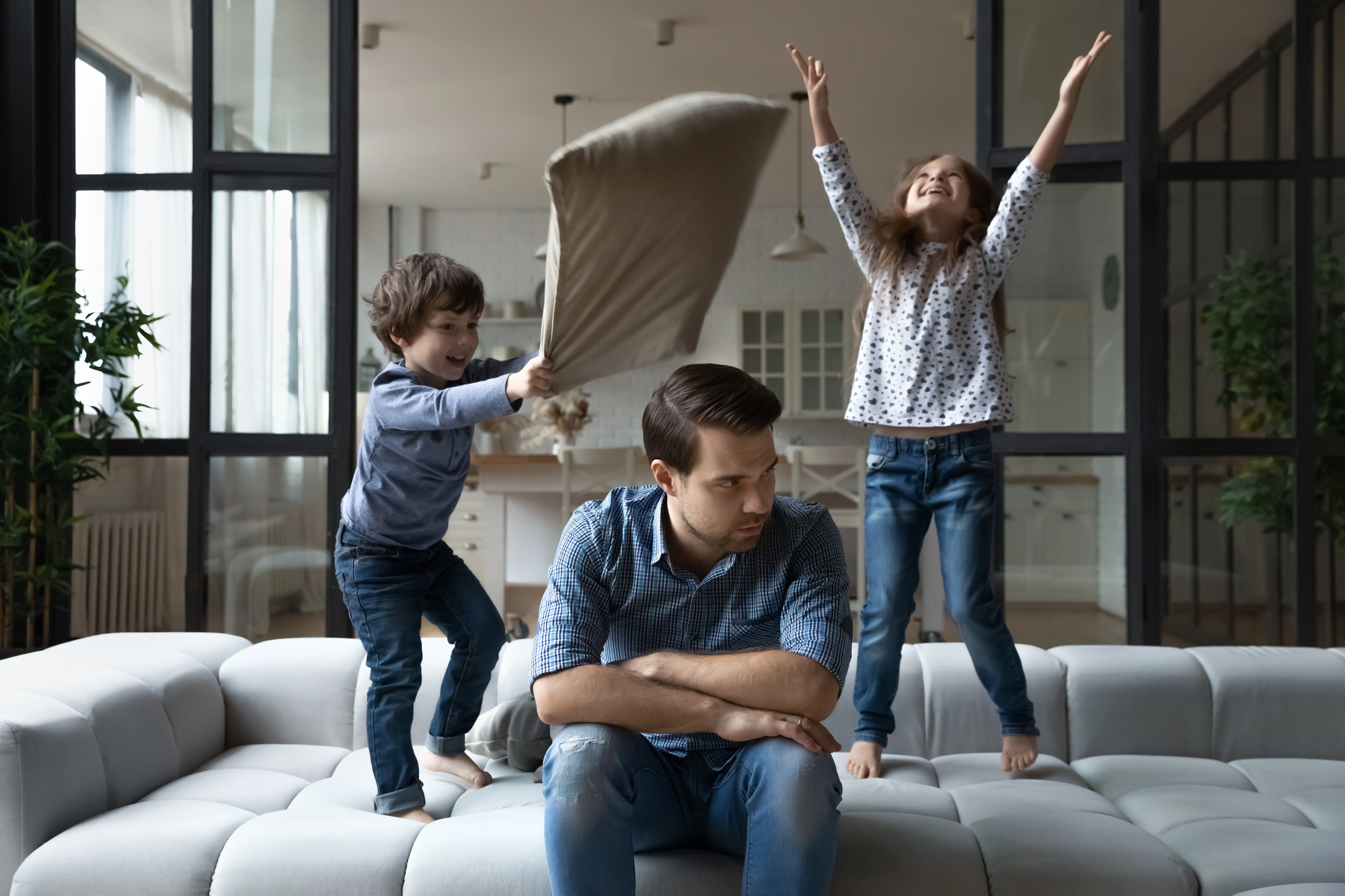 A tired man sits on a couch with his head resting on his hand, while two playful children stand on the couch behind him. One child is raising a pillow, and the other has arms outstretched in excitement. The background shows a modern, open-concept living space.